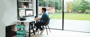 Man sat at desk at home looking at his laptop with folders and files on shelves and a patio door with a garden in the background